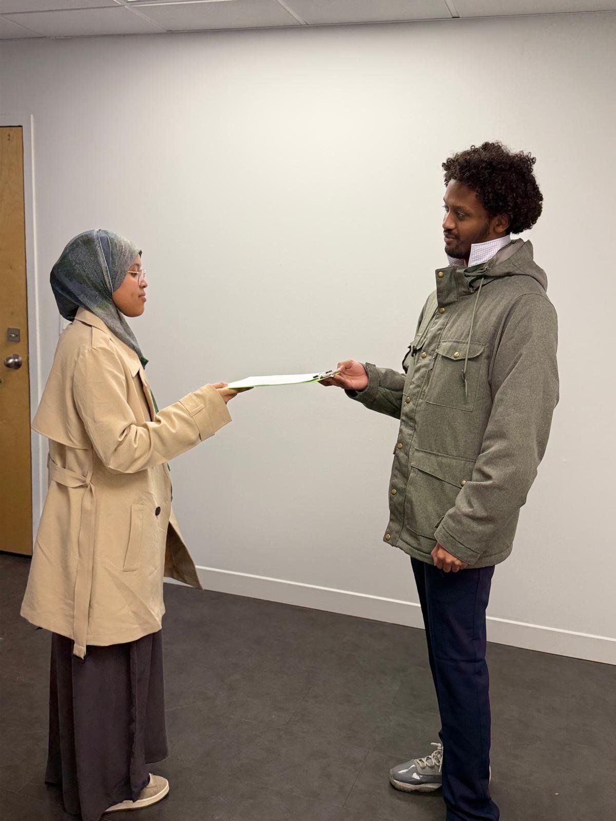 A woman in a headscarf and beige coat hands a document to a man in a green jacket.