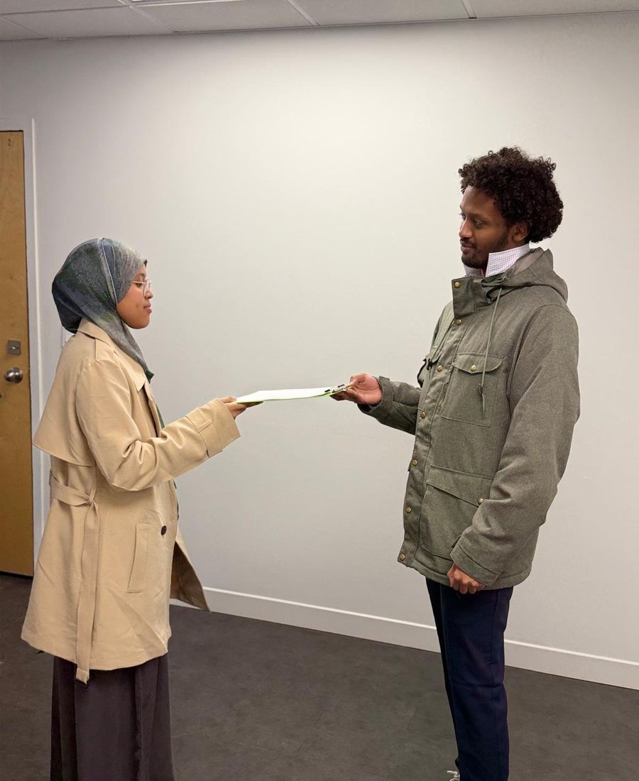 A woman in a headscarf and beige coat hands a document to a man in a green jacket.