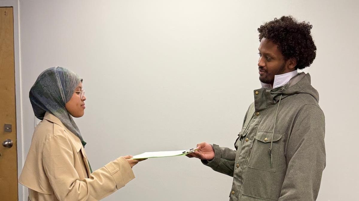 A woman in a headscarf and beige coat hands a document to a man in a green jacket.