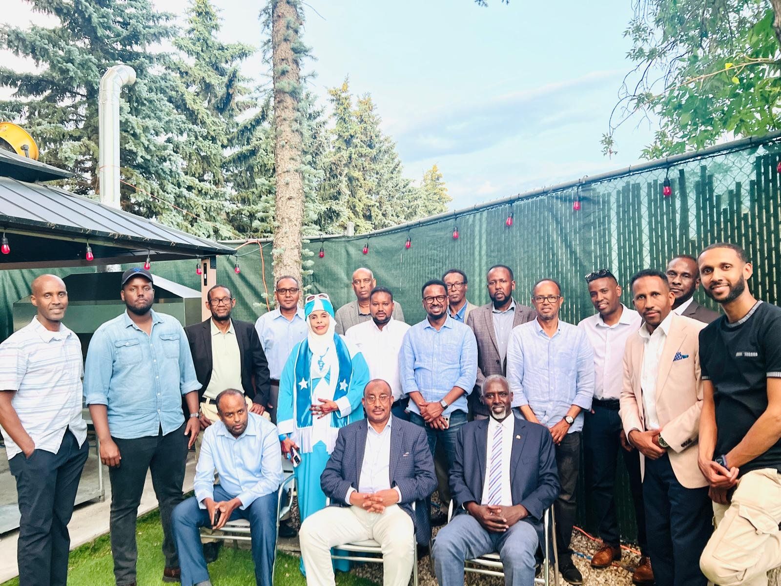 A large group of community members, one wearing a dress in the colours of the Somali flag, poses outdoors among tall trees during Somali Heritage Week.