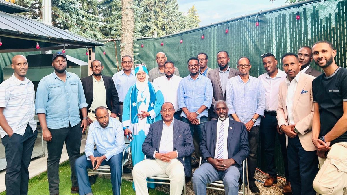 A large group of community members, one wearing a dress in the colours of the Somali flag, poses outdoors among tall trees during Somali Heritage Week.