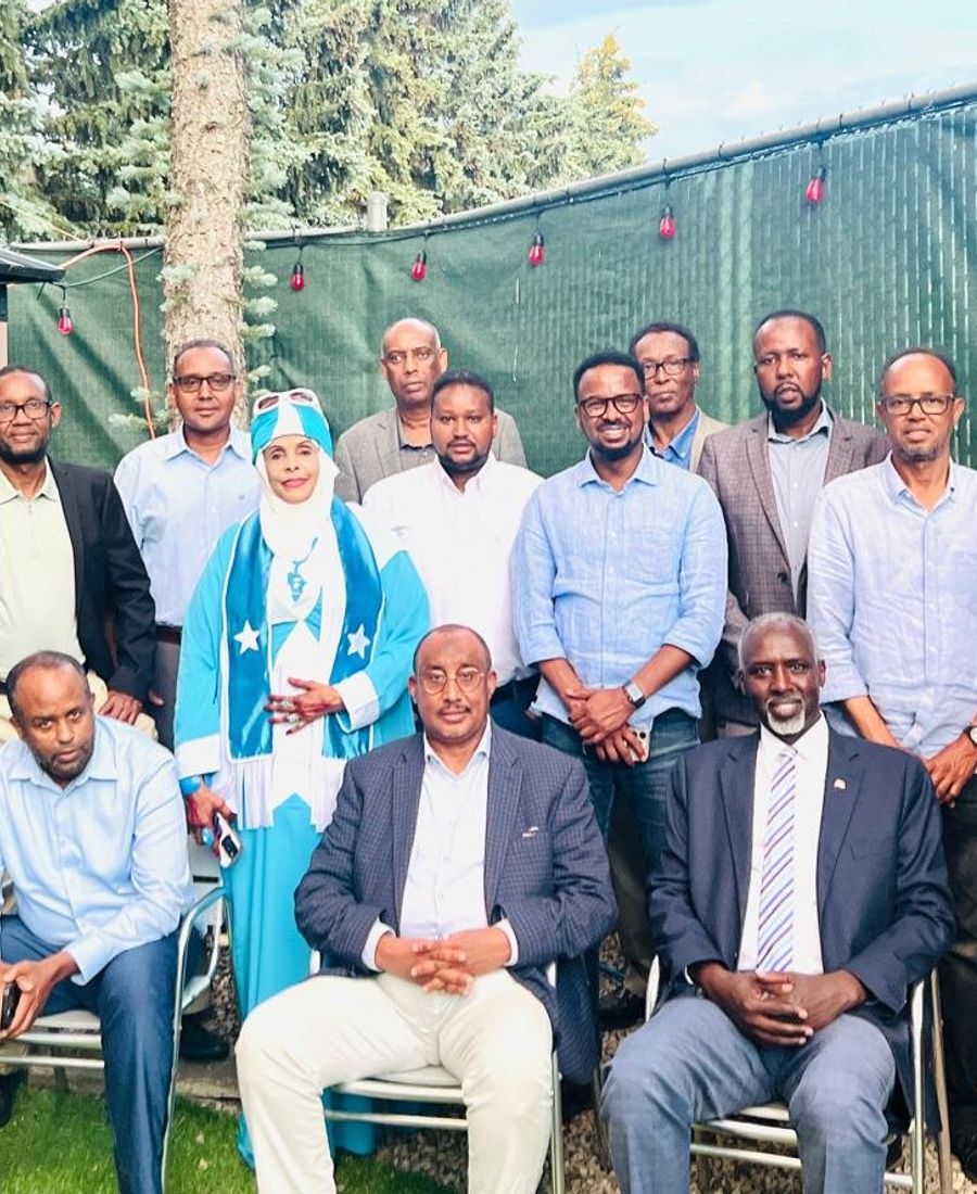 A large group of community members, one wearing a dress in the colours of the Somali flag, poses outdoors among tall trees during Somali Heritage Week.