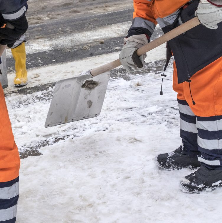 A metal shovel scrapes packed snow and ice off a roadway surface, part of a crew clearing the pavement down to bare ground.