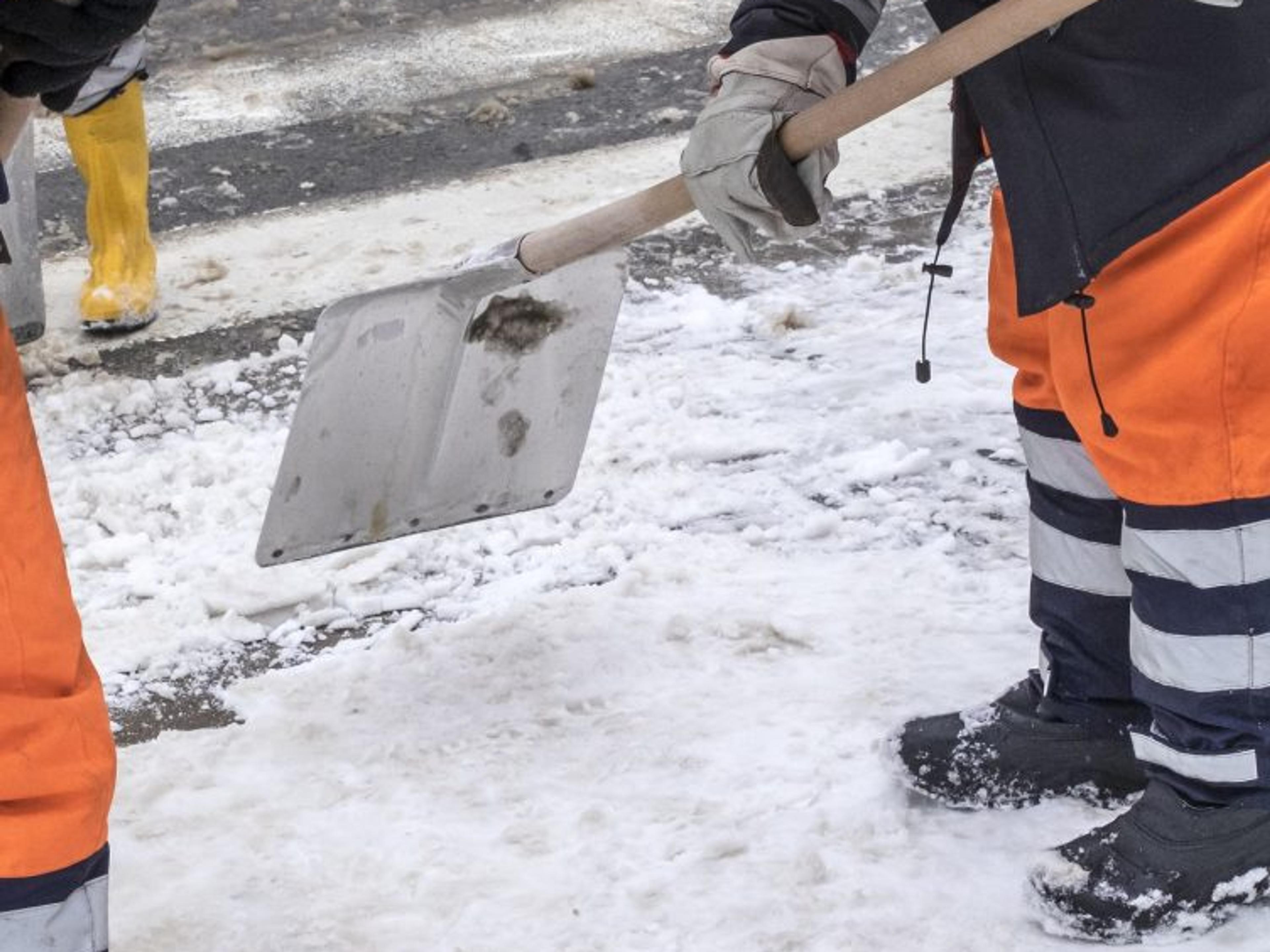 A metal shovel scrapes packed snow and ice off a roadway surface, part of a crew clearing the pavement down to bare ground.