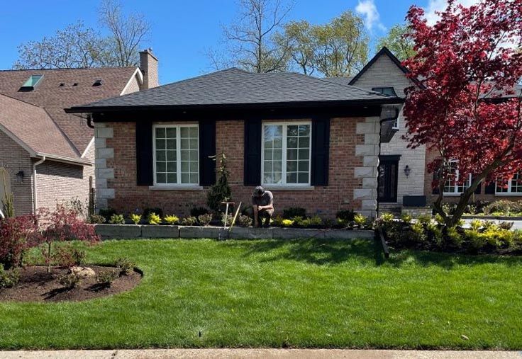 A person tends a garden in front of a brick house, surrounded by lush green grass and a flowering tree on a sunny day.
