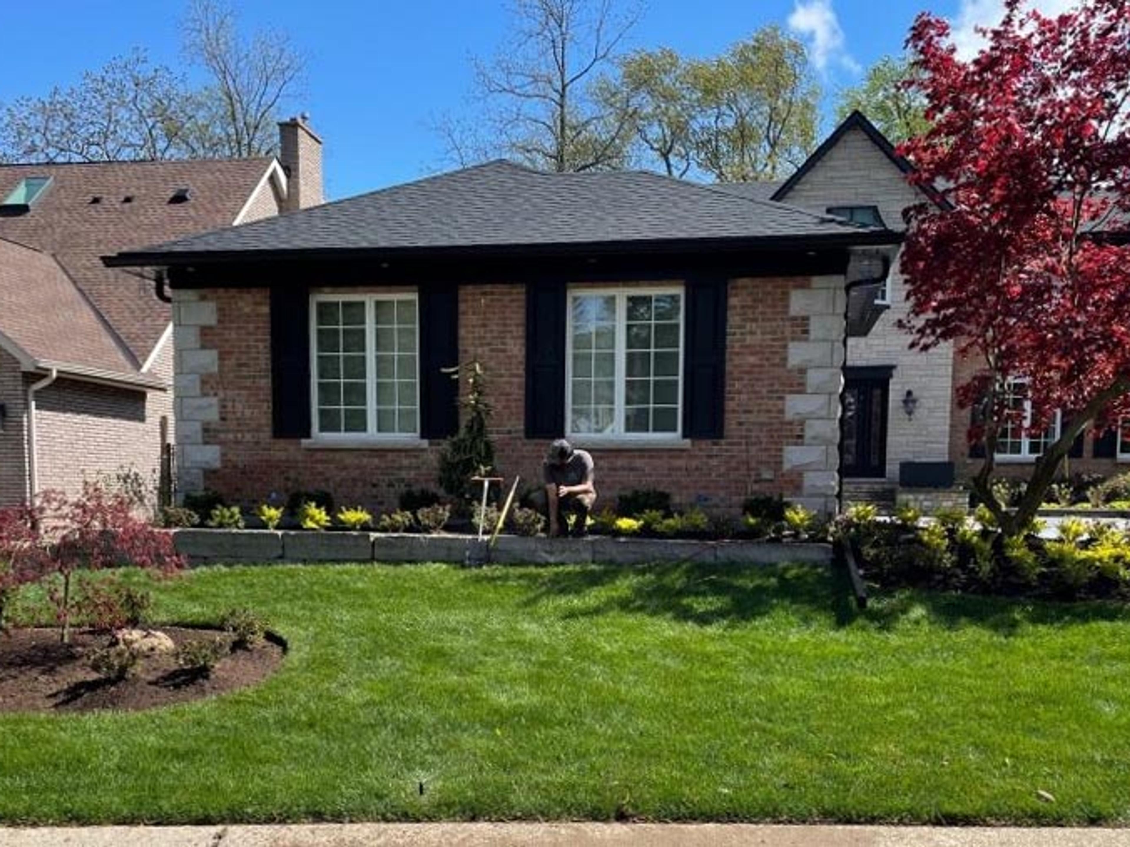 A person tends a garden in front of a brick house, surrounded by lush green grass and a flowering tree on a sunny day.