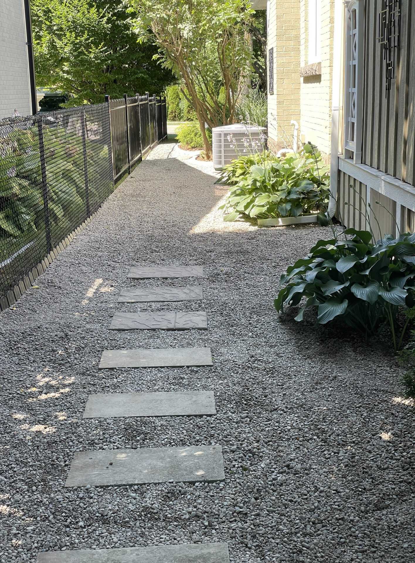 A gravel side path with flagstone stepping pads runs along the house, bordered by a black metal fence and lush hosta plantings.