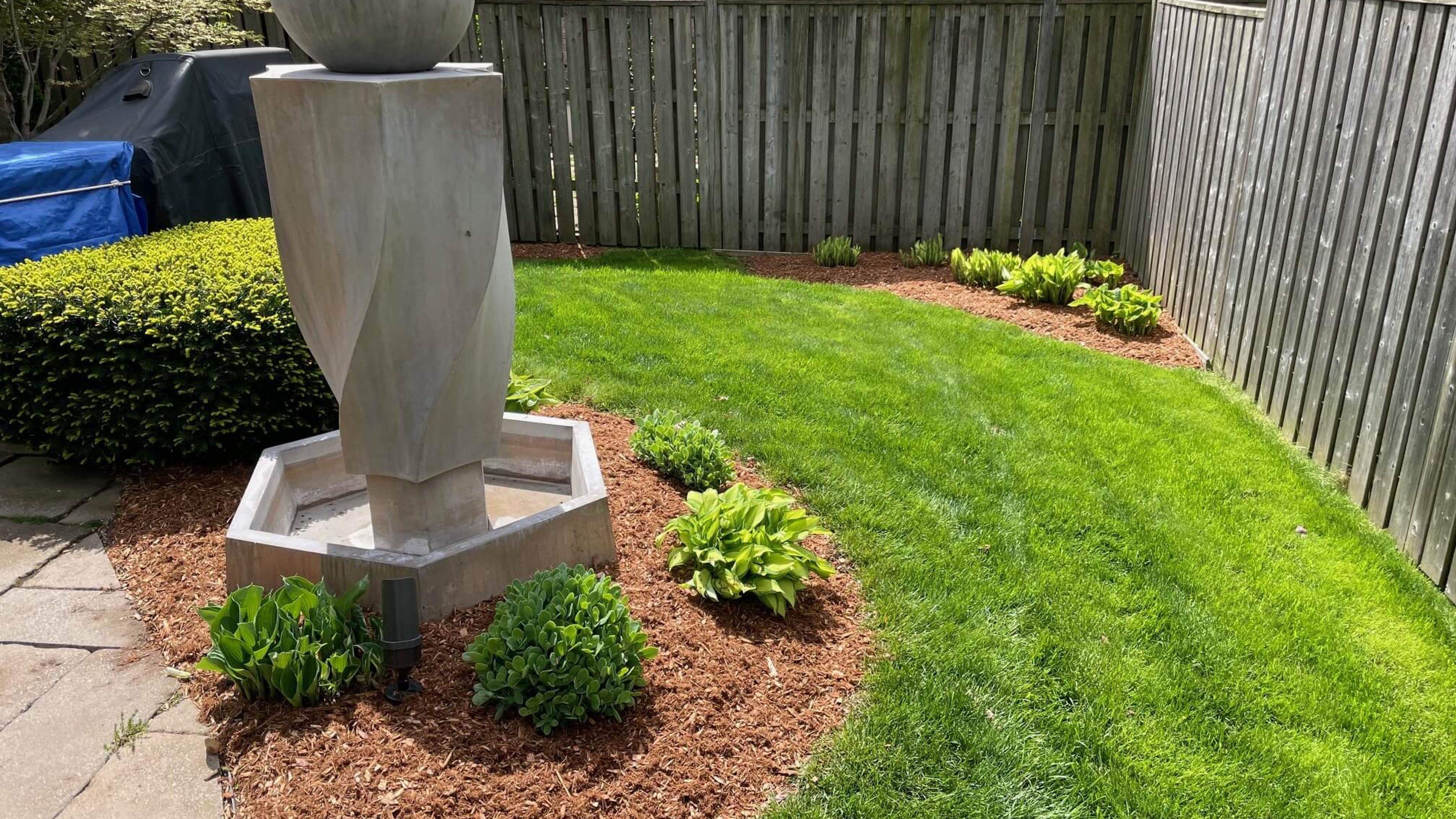 A neatly landscaped garden features a modern sculpture, surrounded by mulch and young plants, enclosed by a wooden fence and patio area.