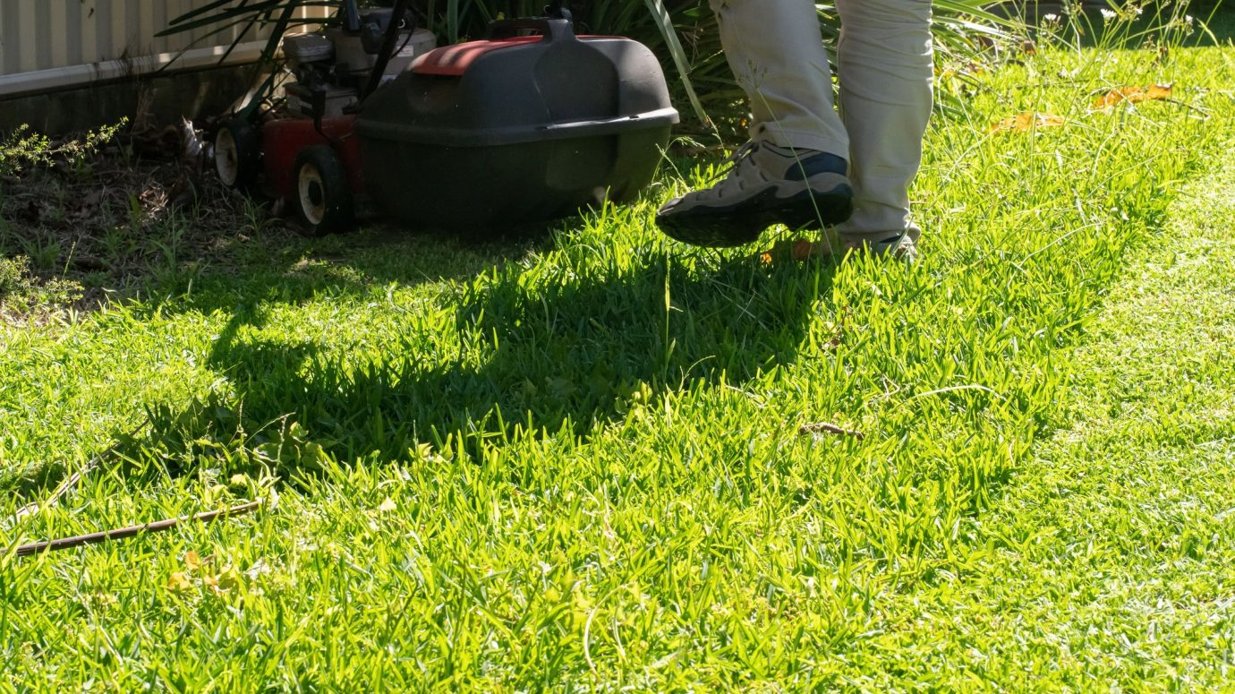 A person mowing lawn with a push mower in a sunny garden, wearing light pants and shoes, next to a metal fence.