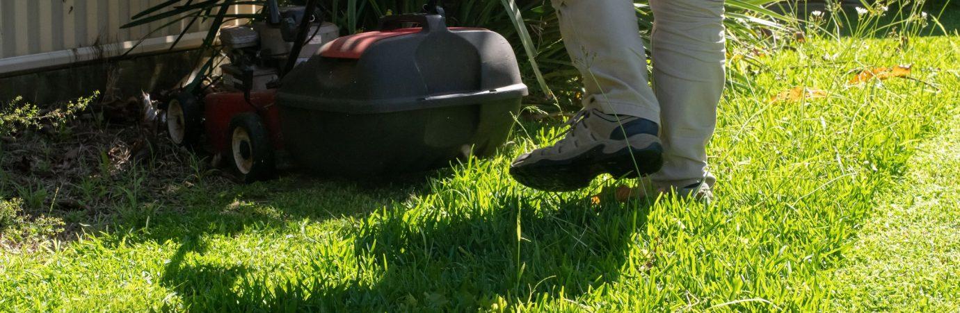 A person mowing lawn with a push mower in a sunny garden, wearing light pants and shoes, next to a metal fence.
