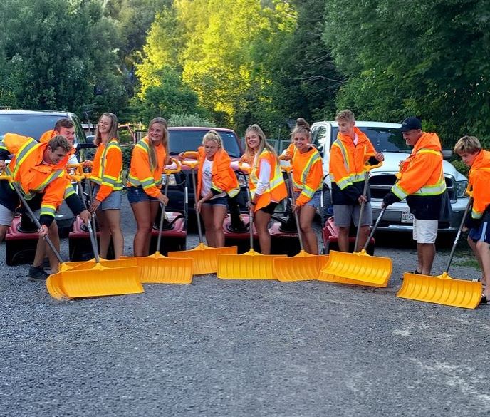 A group of people in fluorescent jackets poses with orange snow shovels in front of trucks, standing on a gravel surface outdoors.