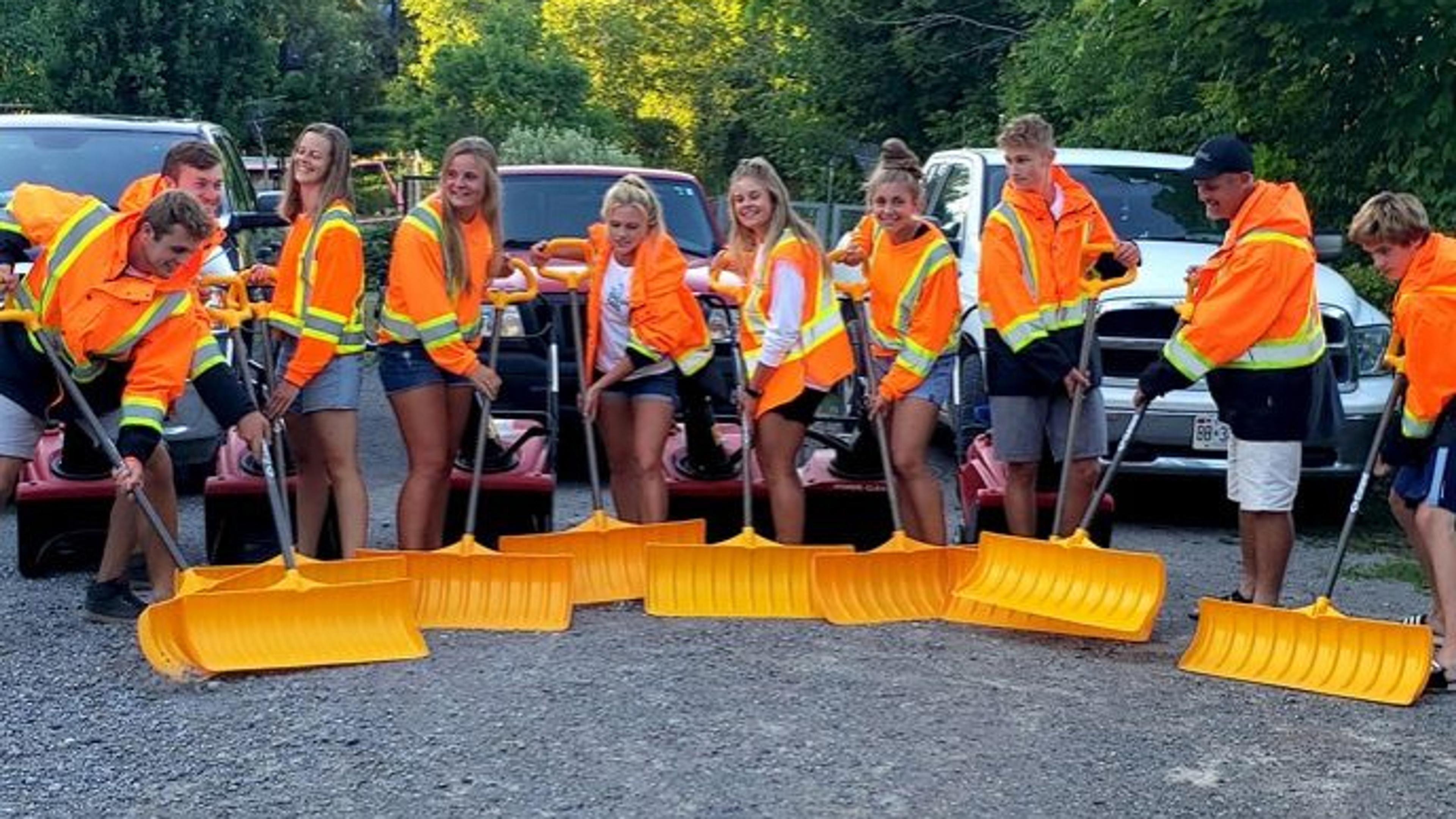 A group of people in fluorescent jackets poses with orange snow shovels in front of trucks, standing on a gravel surface outdoors.