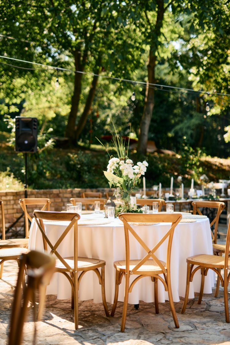 An outdoor event setup with linen covered tables, cross back chairs, and floral centerpieces arranged on a stone patio beneath a wooded backdrop.