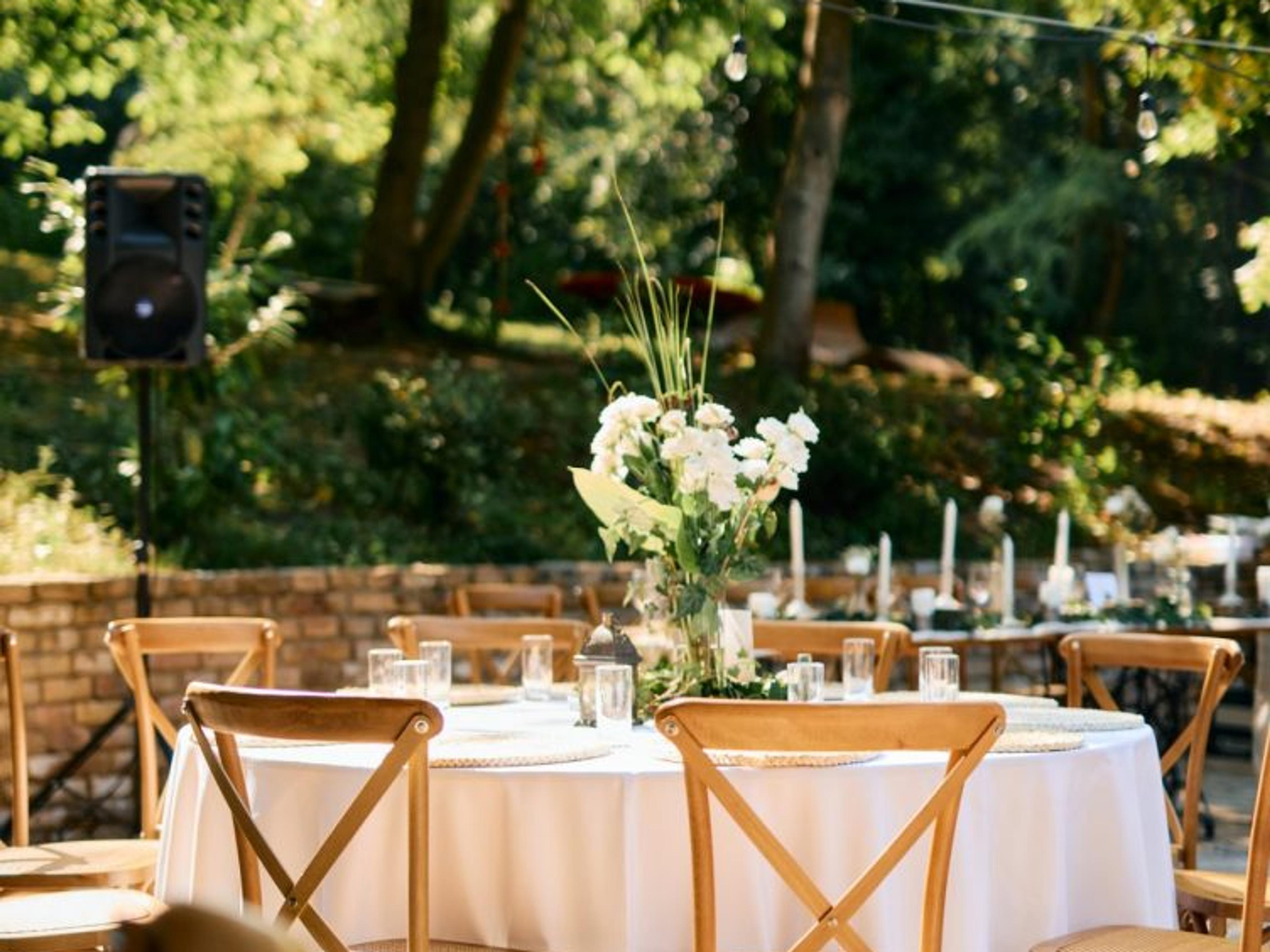 An outdoor event setup with linen covered tables, cross back chairs, and floral centerpieces arranged on a stone patio beneath a wooded backdrop.