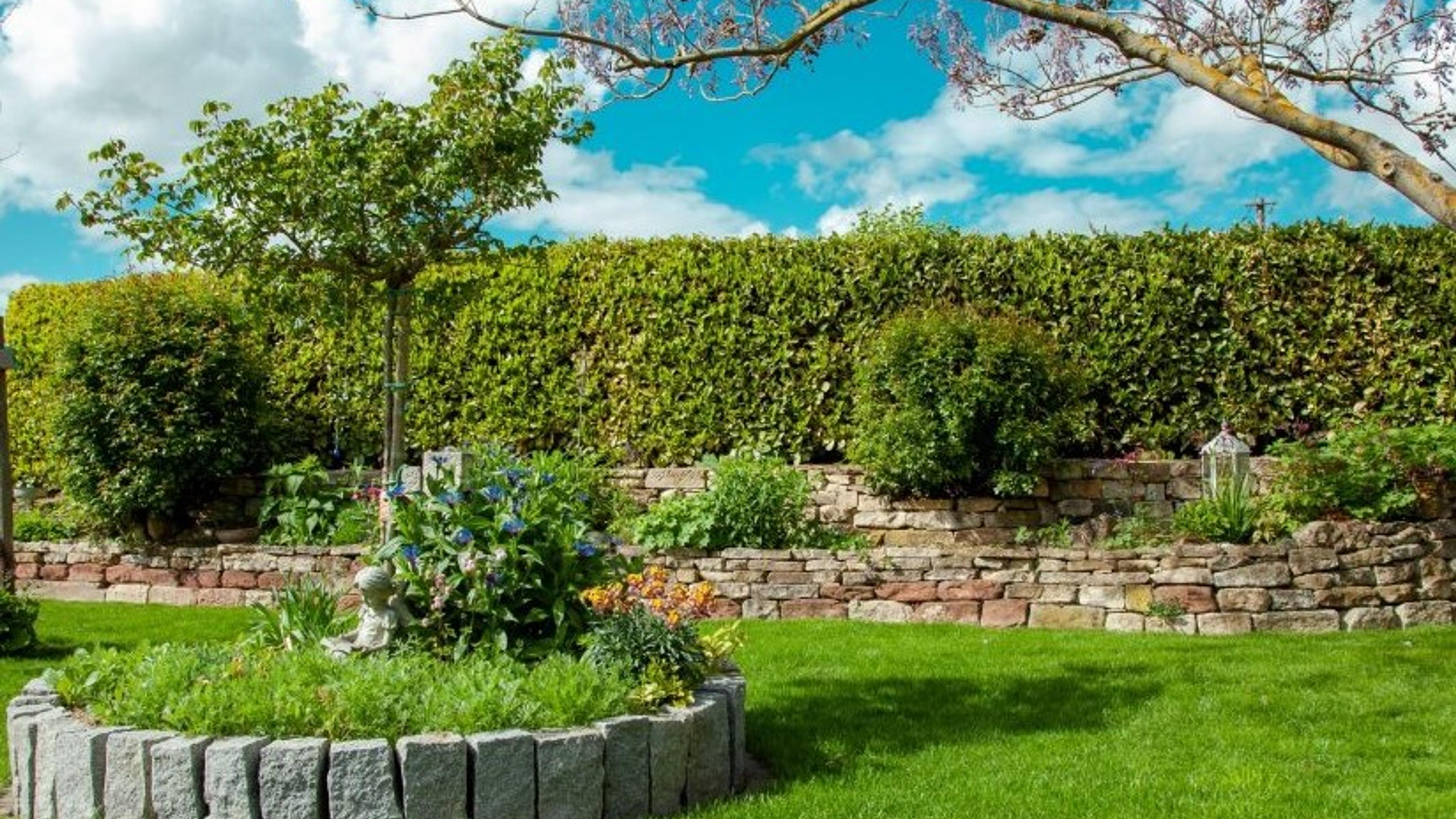 A stone raised planter bursting with hydrangeas and marigolds sits before a rustic stacked stone retaining wall and a tall, tightly clipped hedge, with a manicured lawn rolling out in front.