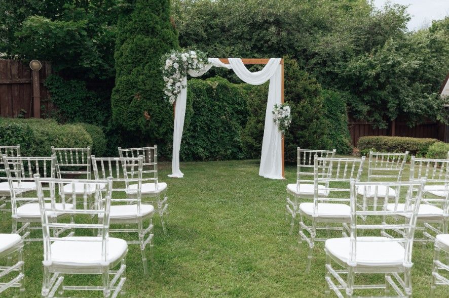 A backyard wedding ceremony setup on a manicured lawn, with clear chiavari chairs facing a floral wooden arch framed by a dense evergreen hedge.
