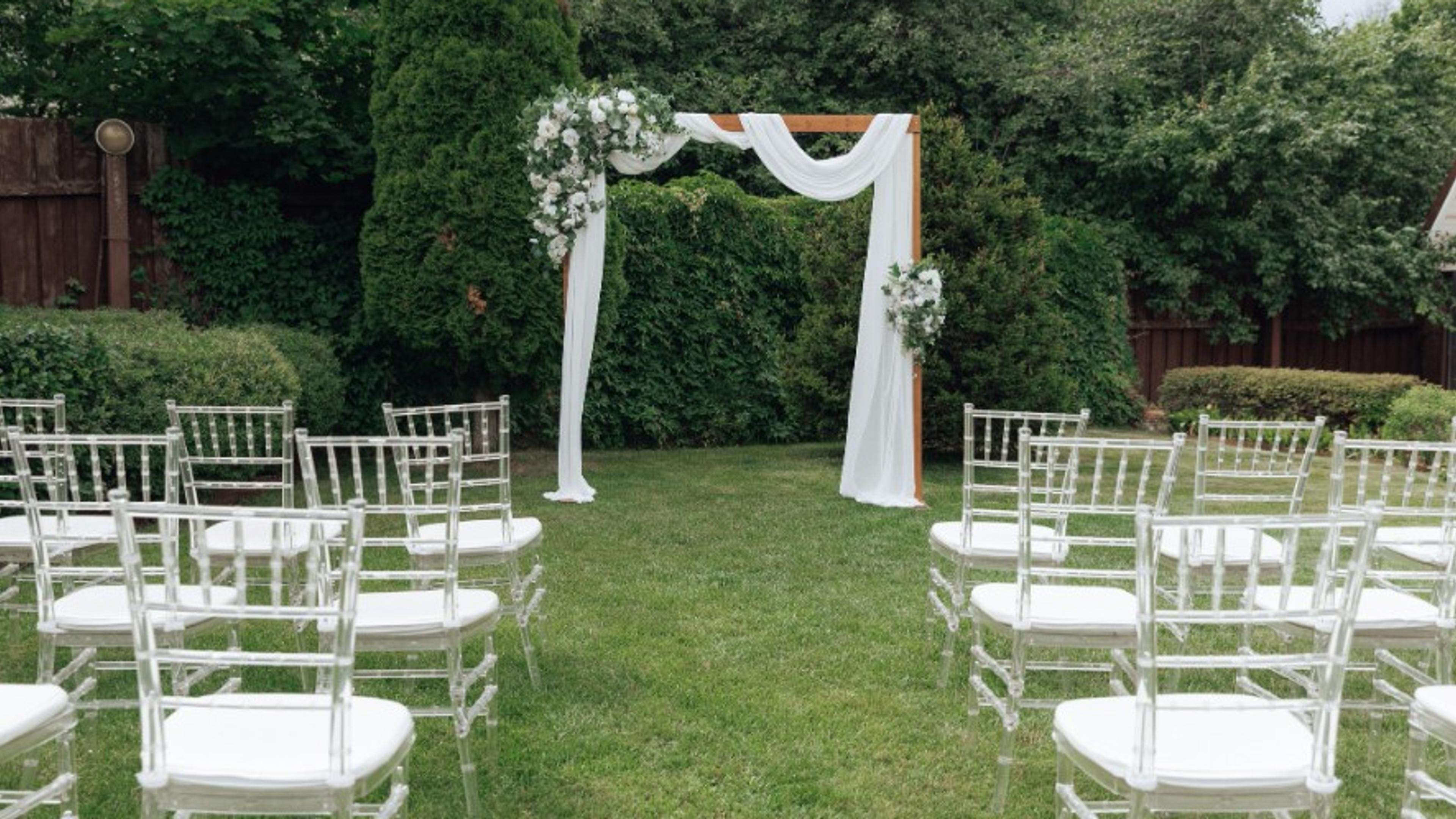 A backyard wedding ceremony setup on a manicured lawn, with clear chiavari chairs facing a floral wooden arch framed by a dense evergreen hedge.