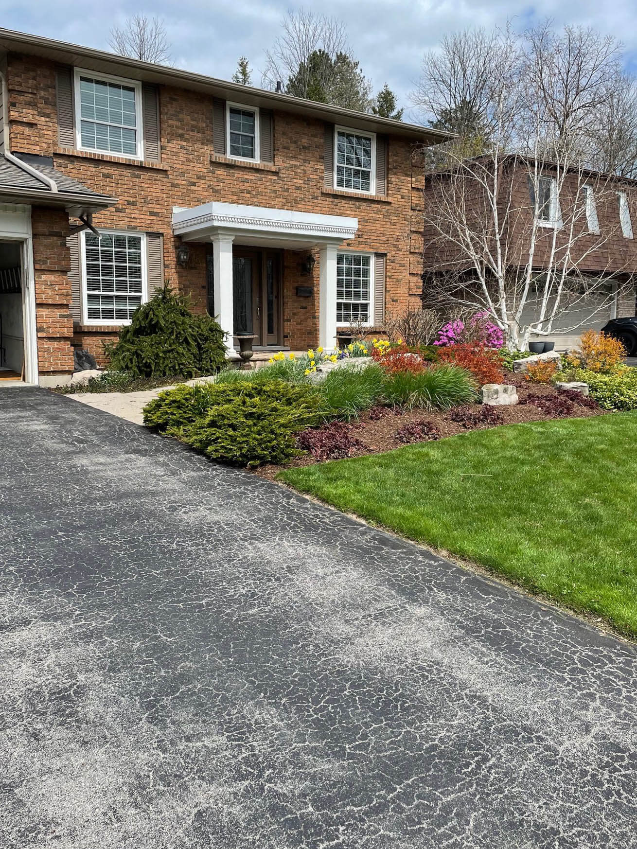 A two-story brick house with a manicured garden, colorful flowers, and a driveway. No recognizable landmarks or historical buildings are visible.