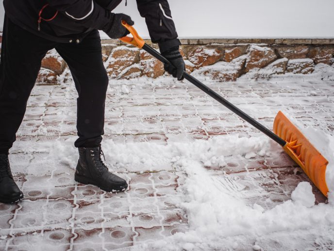 A snow shovel clears fresh snow off a paver walkway, revealing the clean patterned brick surface beneath.