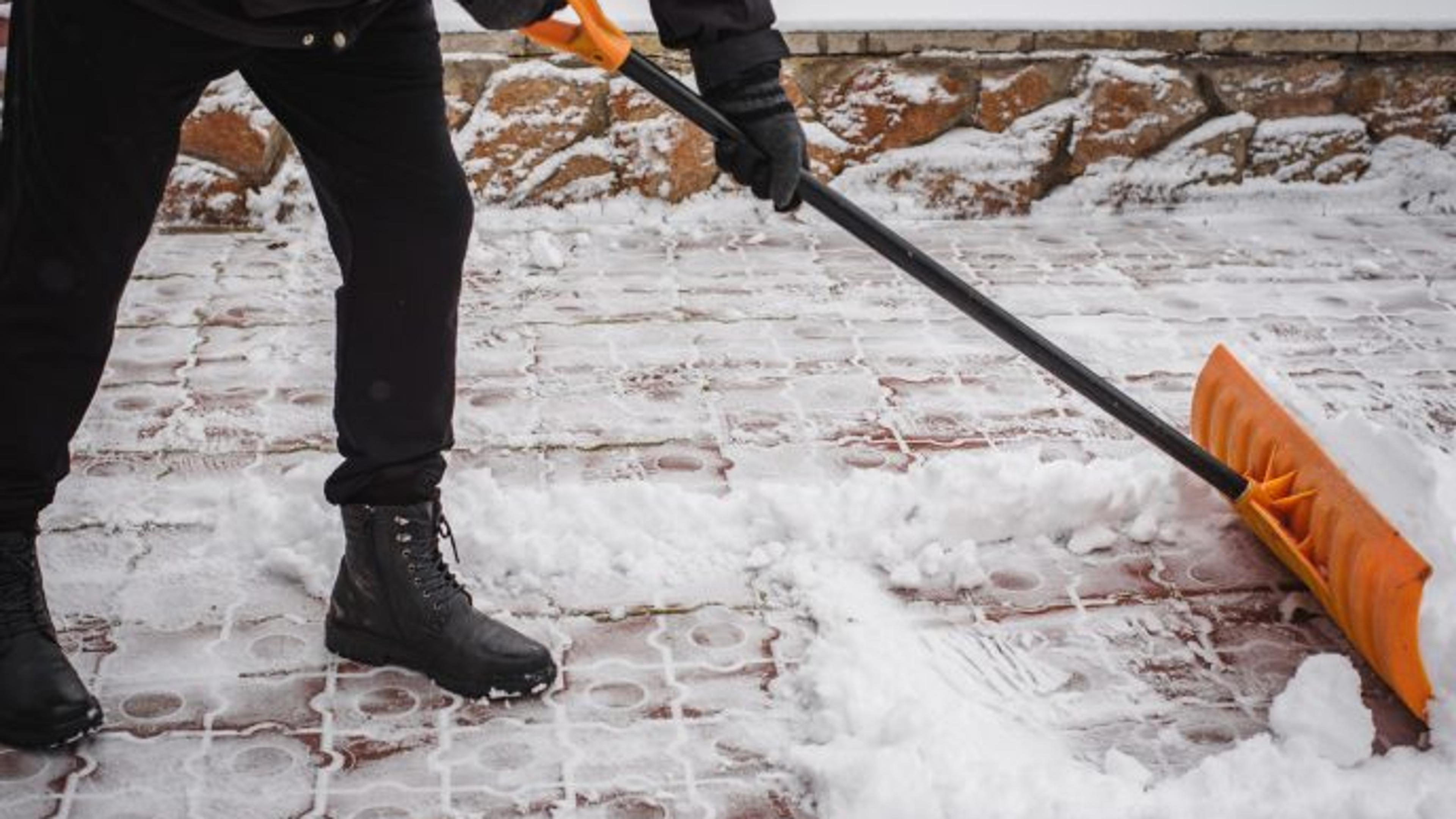 A snow shovel clears fresh snow off a paver walkway, revealing the clean patterned brick surface beneath.