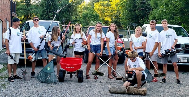 A group of people in white shirts with garden tools stand in front of vehicles; a Chapin red wheelbarrow is in the center.