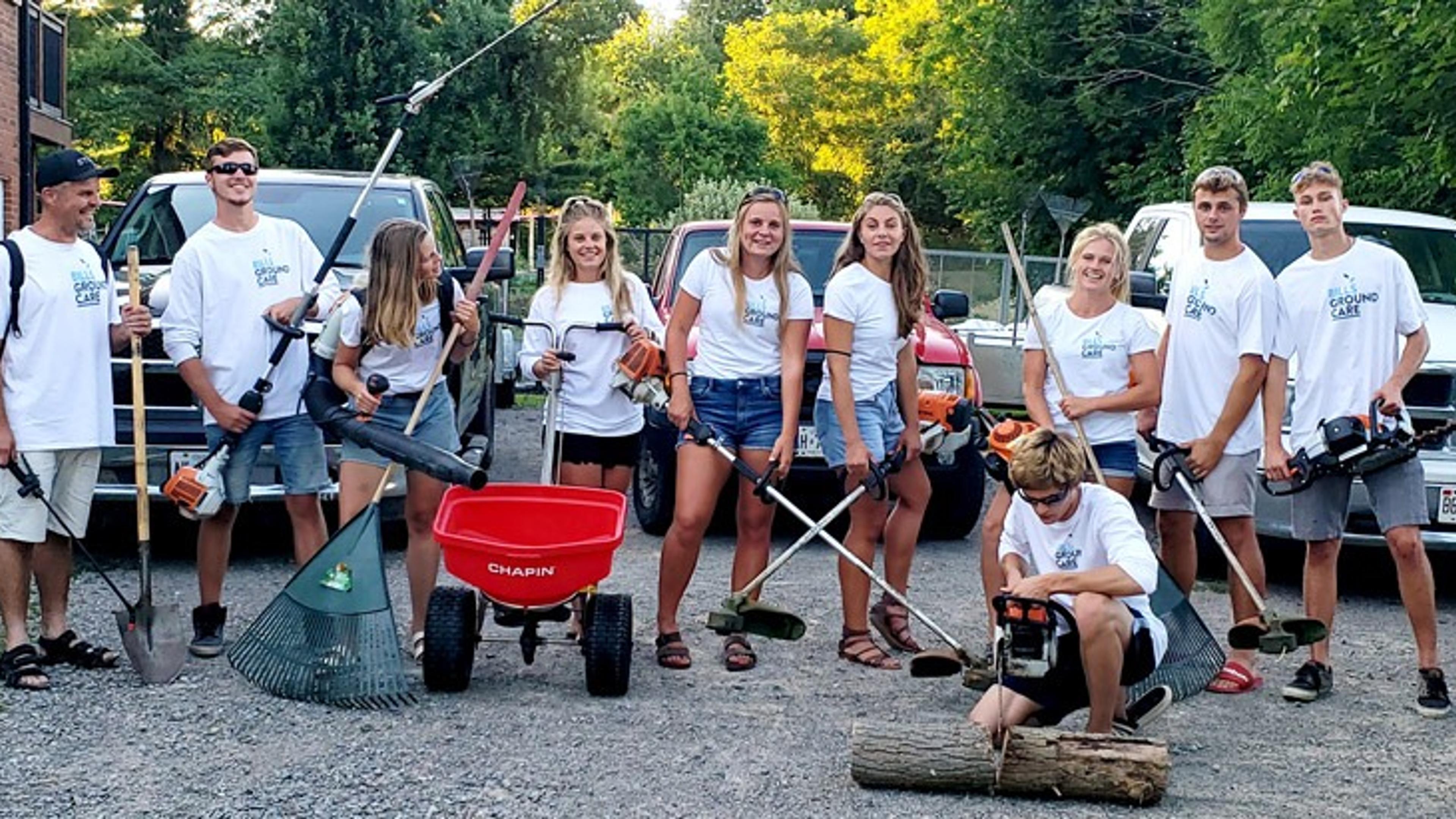 A group of people in white shirts with garden tools stand in front of vehicles; a Chapin red wheelbarrow is in the center.