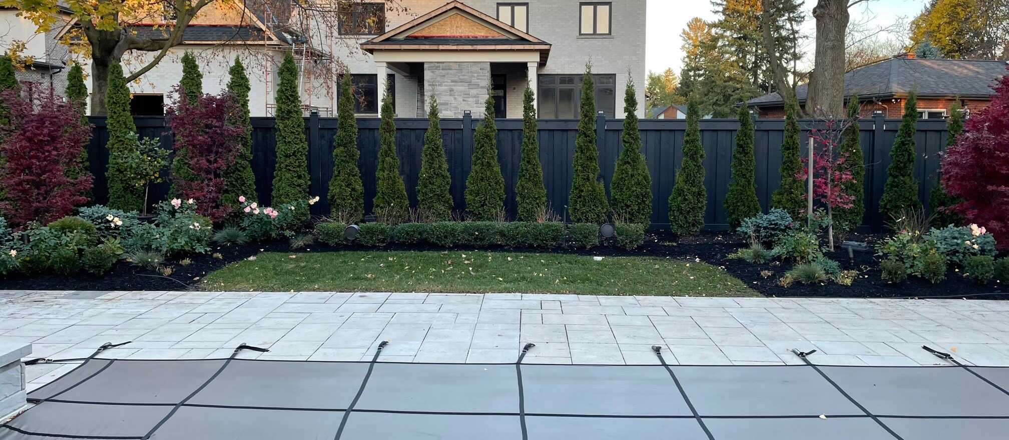 A modern backyard features a covered pool, stone patio, manicured lawn, tall evergreen trees, vibrant shrubs, and a gray house in the background.