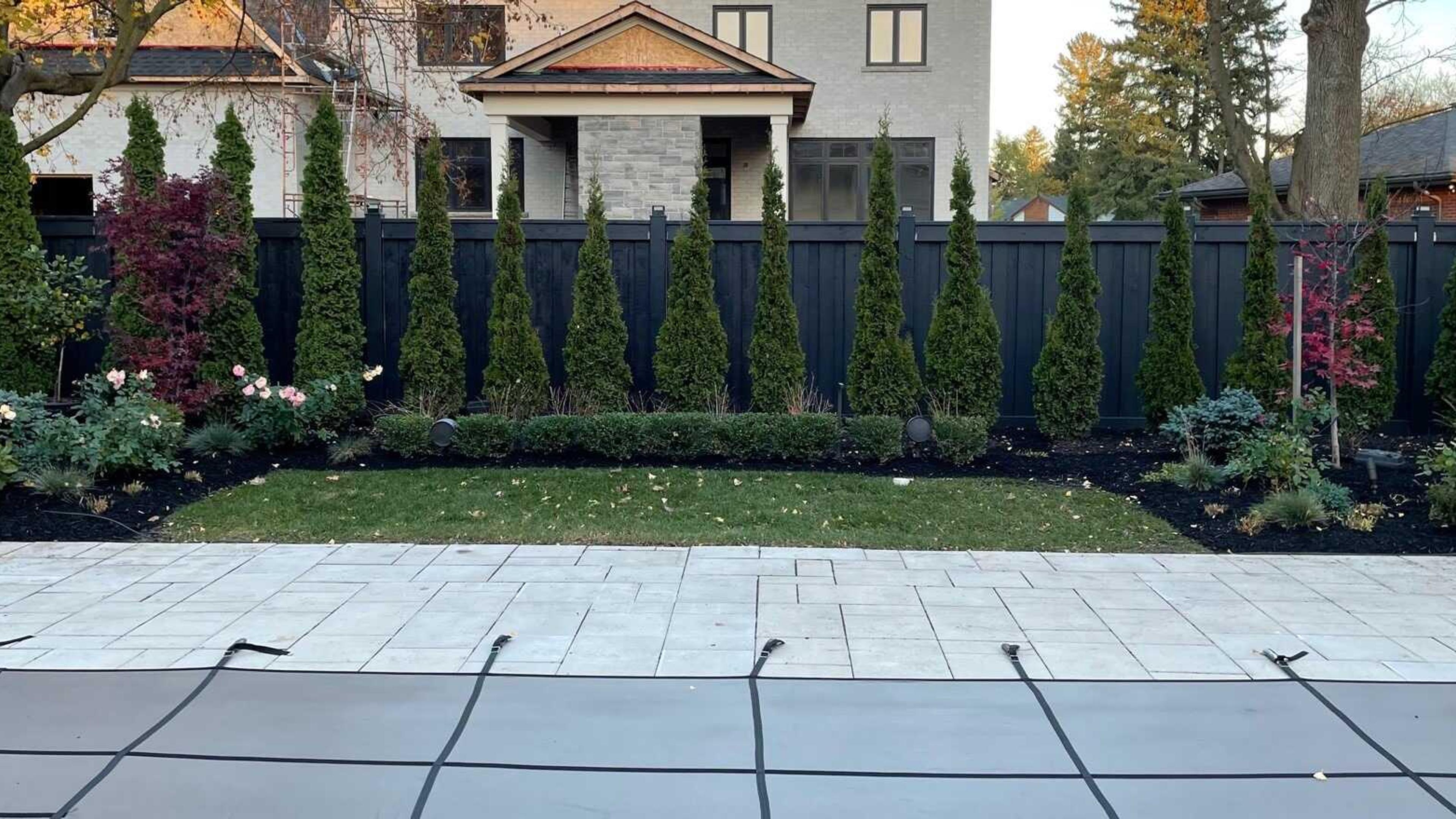 A modern backyard features a covered pool, stone patio, manicured lawn, tall evergreen trees, vibrant shrubs, and a gray house in the background.