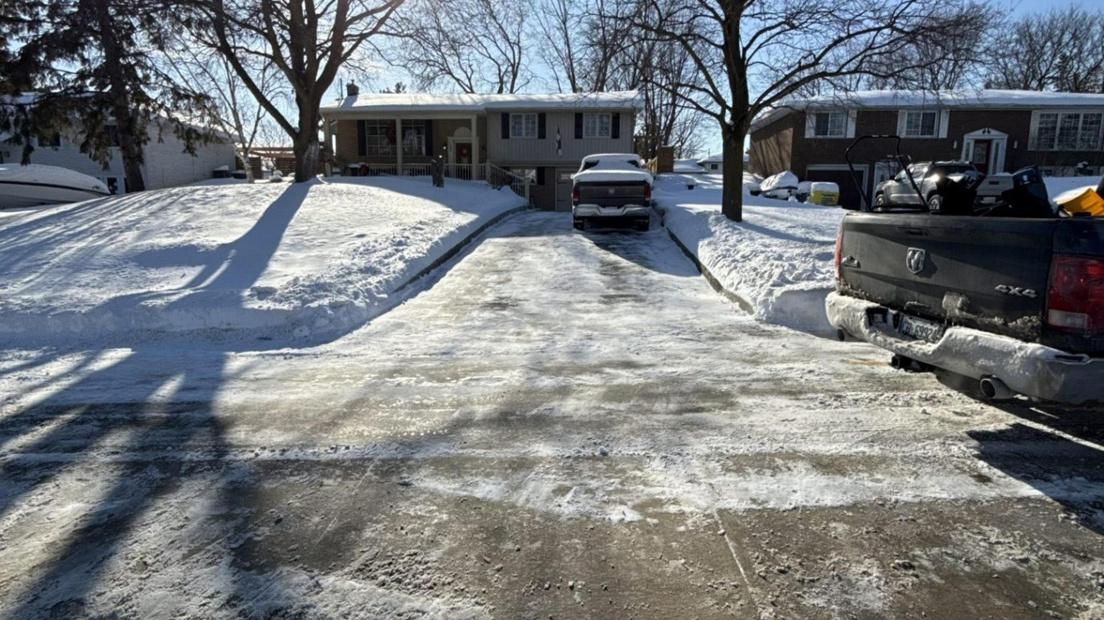 A residential driveway freshly cleared of snow and ice down to bare pavement, with the surrounding lawn and walkways still snow covered.