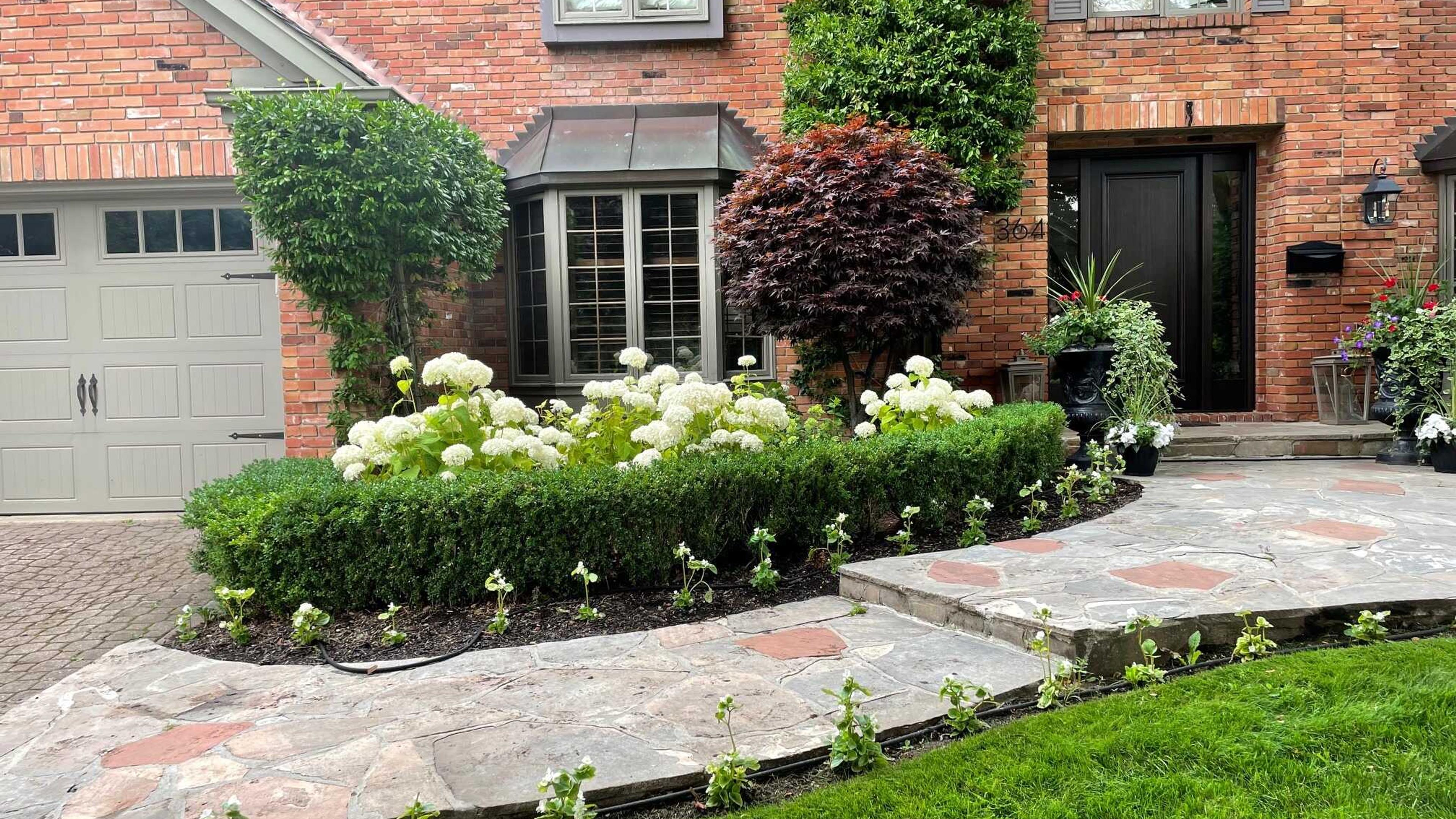 Brick house with well-manicured garden, stone pathway, and various potted plants. Features a two-car garage and a bay window.