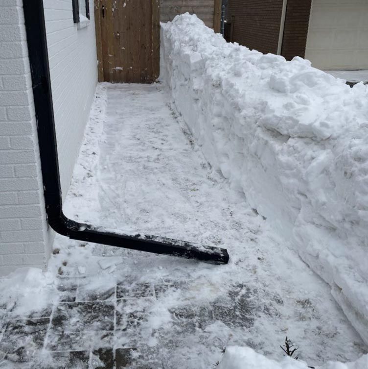 Narrow pathway between two houses with high snow piles. Snow-covered ground drains into a black gutter pipe attached to the wall.