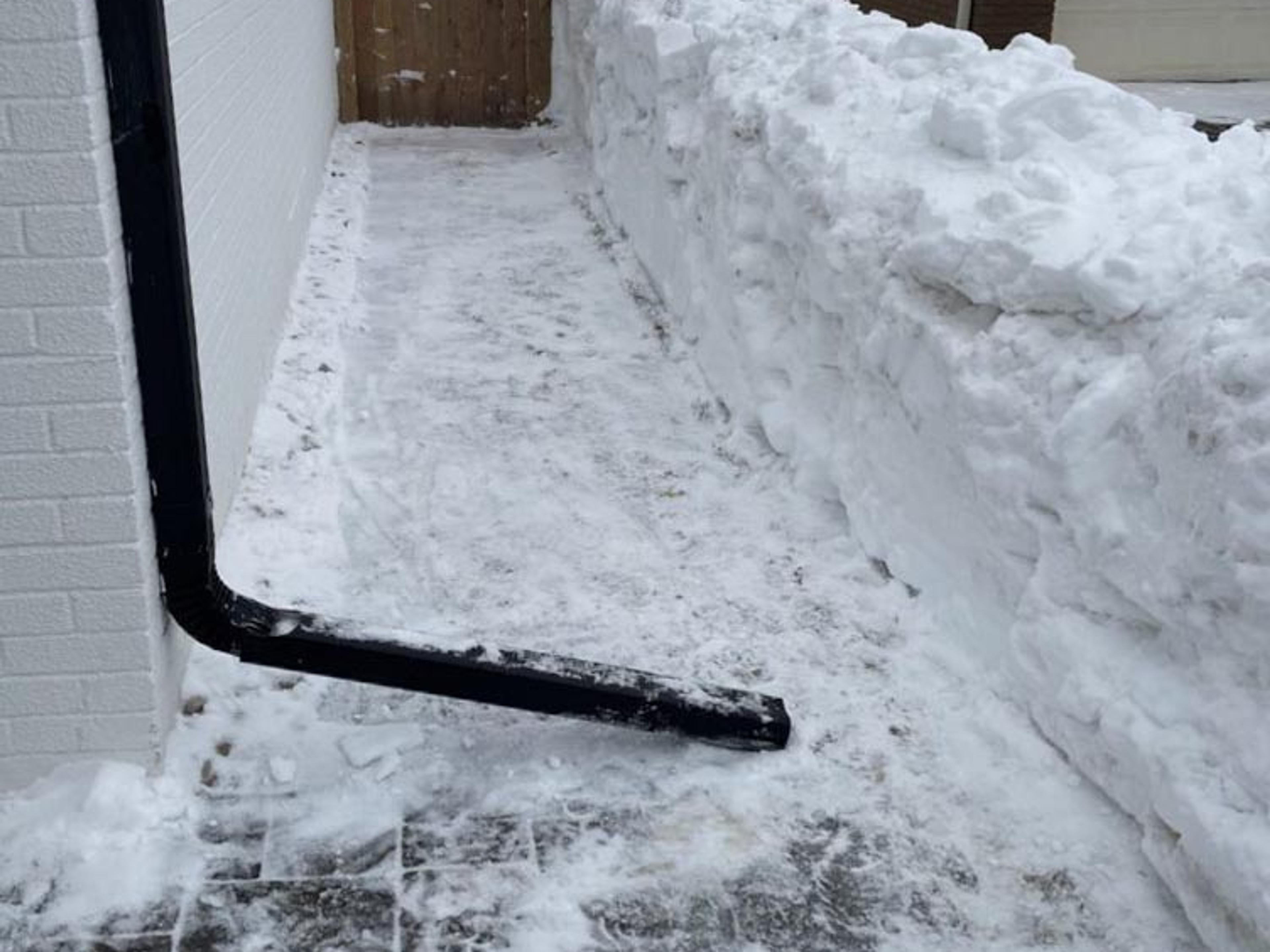 Narrow pathway between two houses with high snow piles. Snow-covered ground drains into a black gutter pipe attached to the wall.