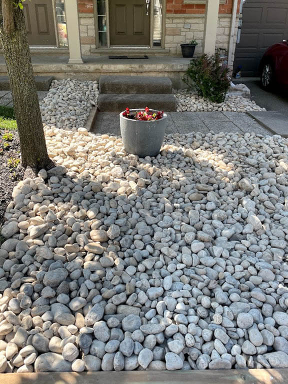 A front yard with pebble landscaping, a tree, potted flowers, and a red car parked near a brick house entrance with steps.
