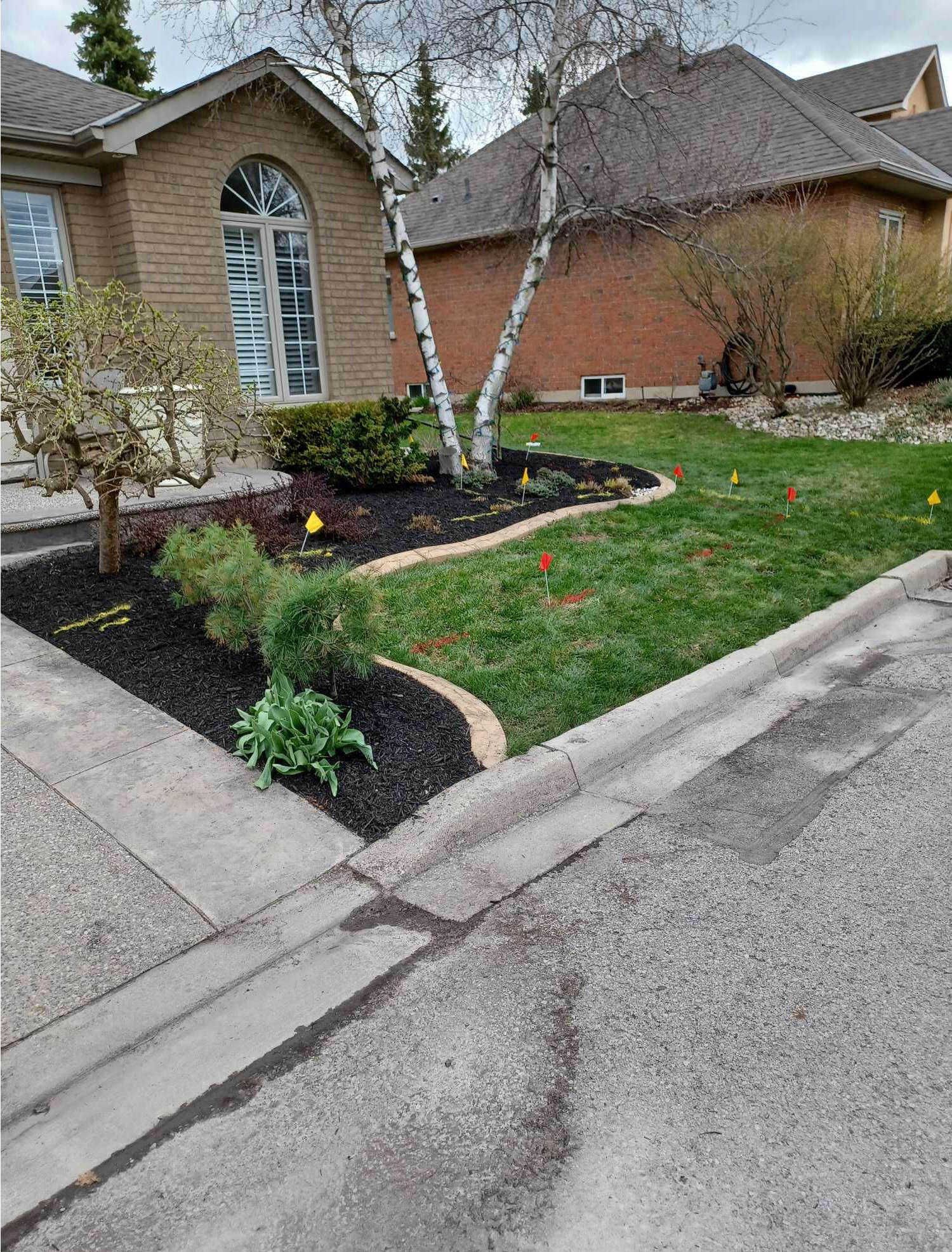A suburban house with manicured garden, featuring mulch, small trees, and colorful markers on the lawn. No recognizable landmarks or historical buildings.