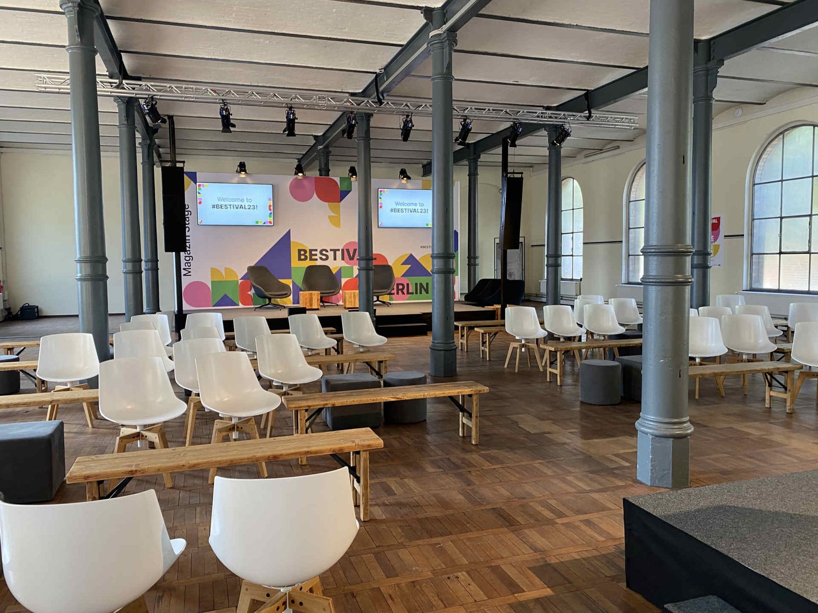 Industrielle Venue mit Säulen, Holzboden, heller Bestuhlung und bunter Pressewand im Hintergrund