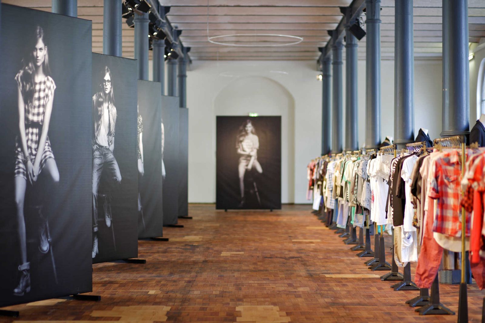 Long hall with wooden parquet flooring and a long row of clothing racks on the right and large banners with fashion photography on the left.