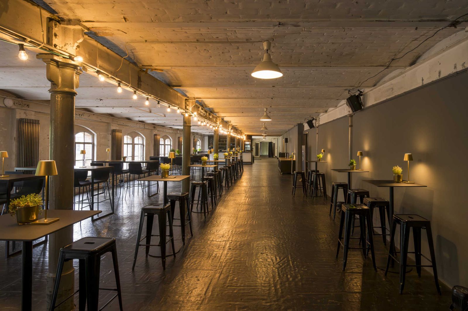 Langes industrielles Loft mit vielen schwarzen Tischen und goldenen Lampen.