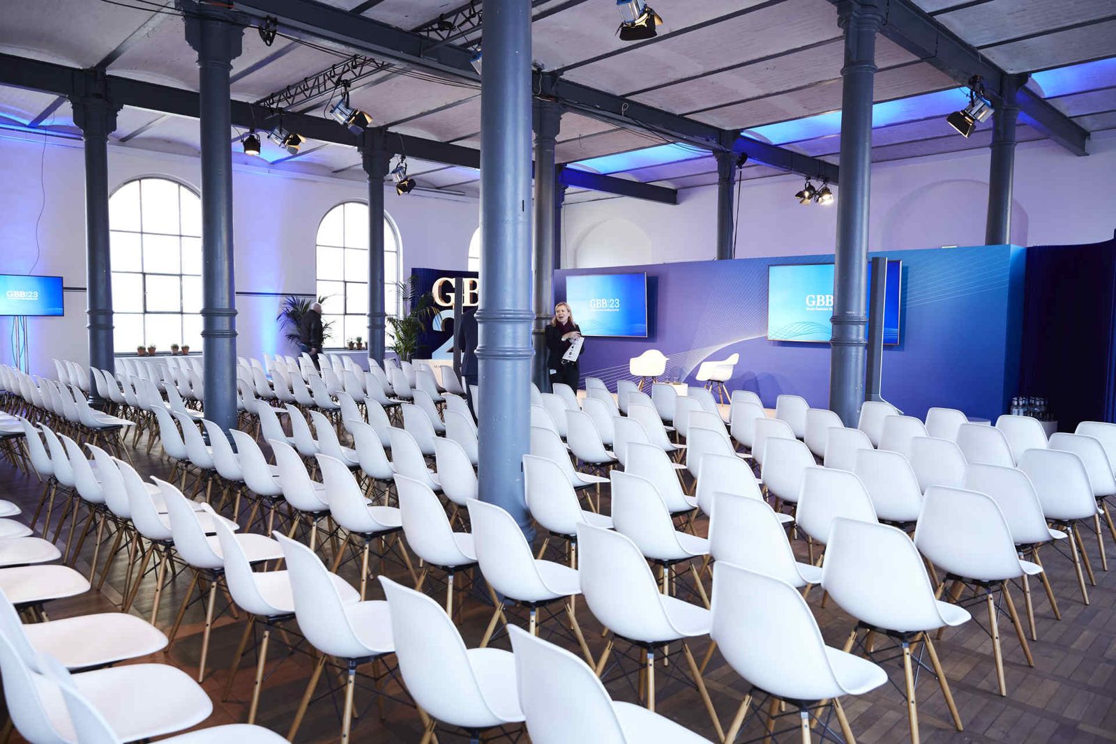 weiße Reihenbestuhlung vor einer Bühne mit großem blauen Backdrop mit Screens