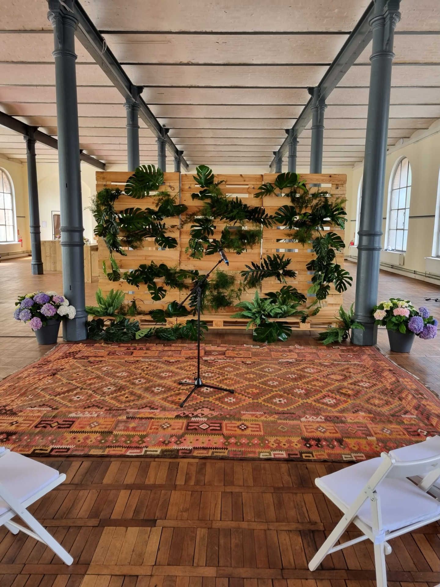 Großer Saal mit Zeremonie Setting für Trauung vor begrünter Palettenwand.