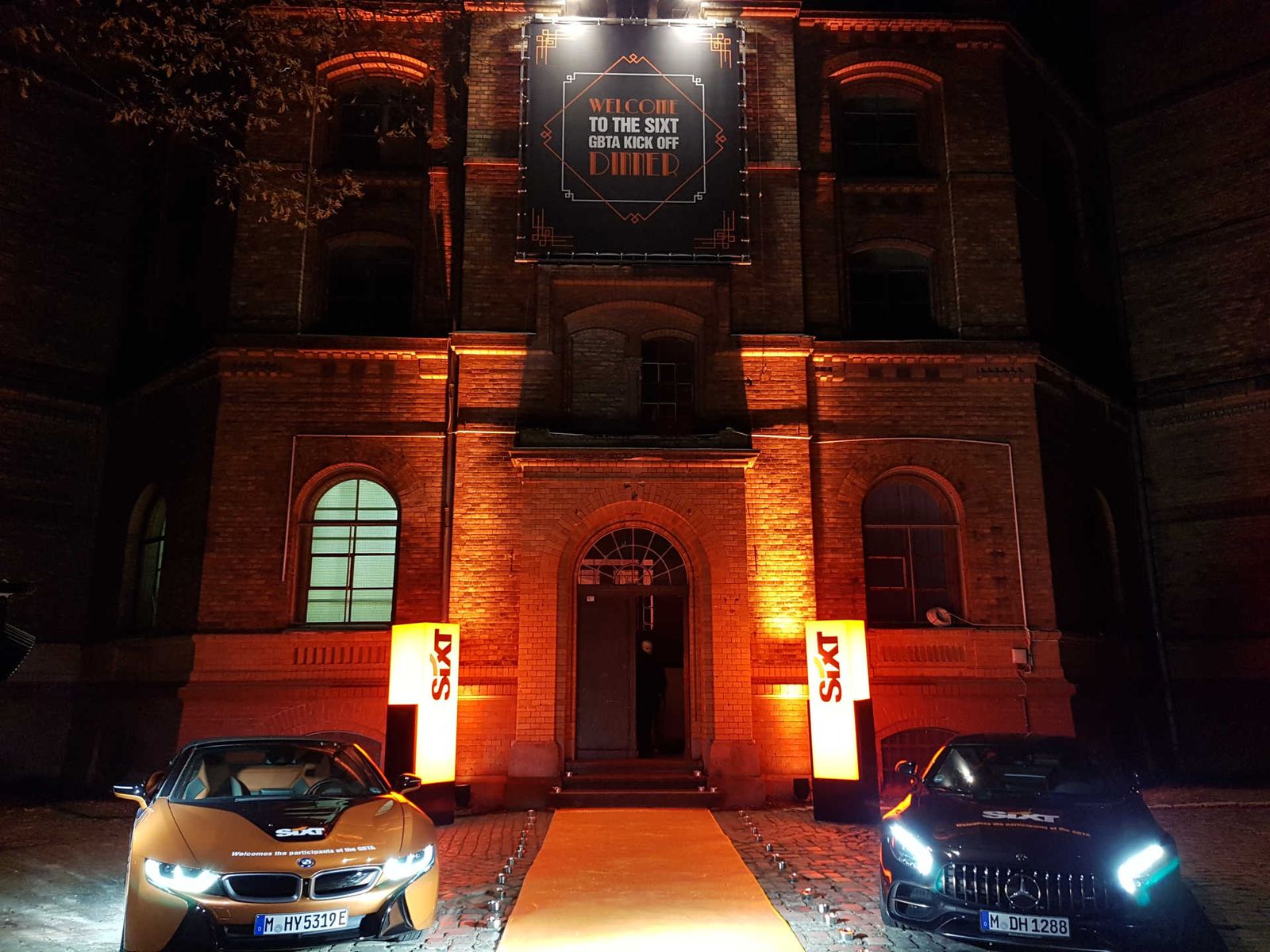 Main entrance of large brick building with two showcased cars under dramatic lighting.