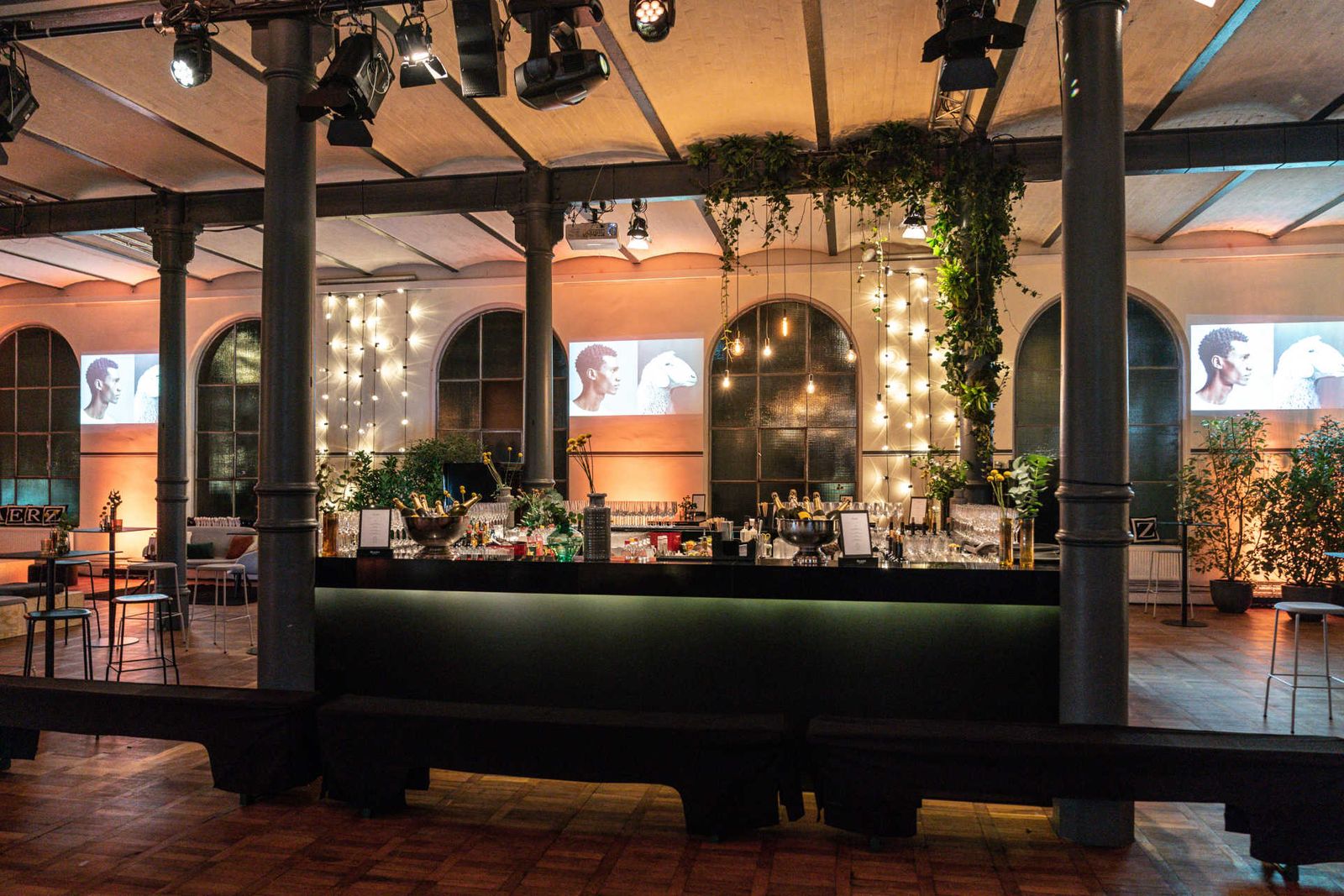 Atmosphärisch beleuchtete Eventlocation mit zentraler Bar und Pflanzen Dekoration.