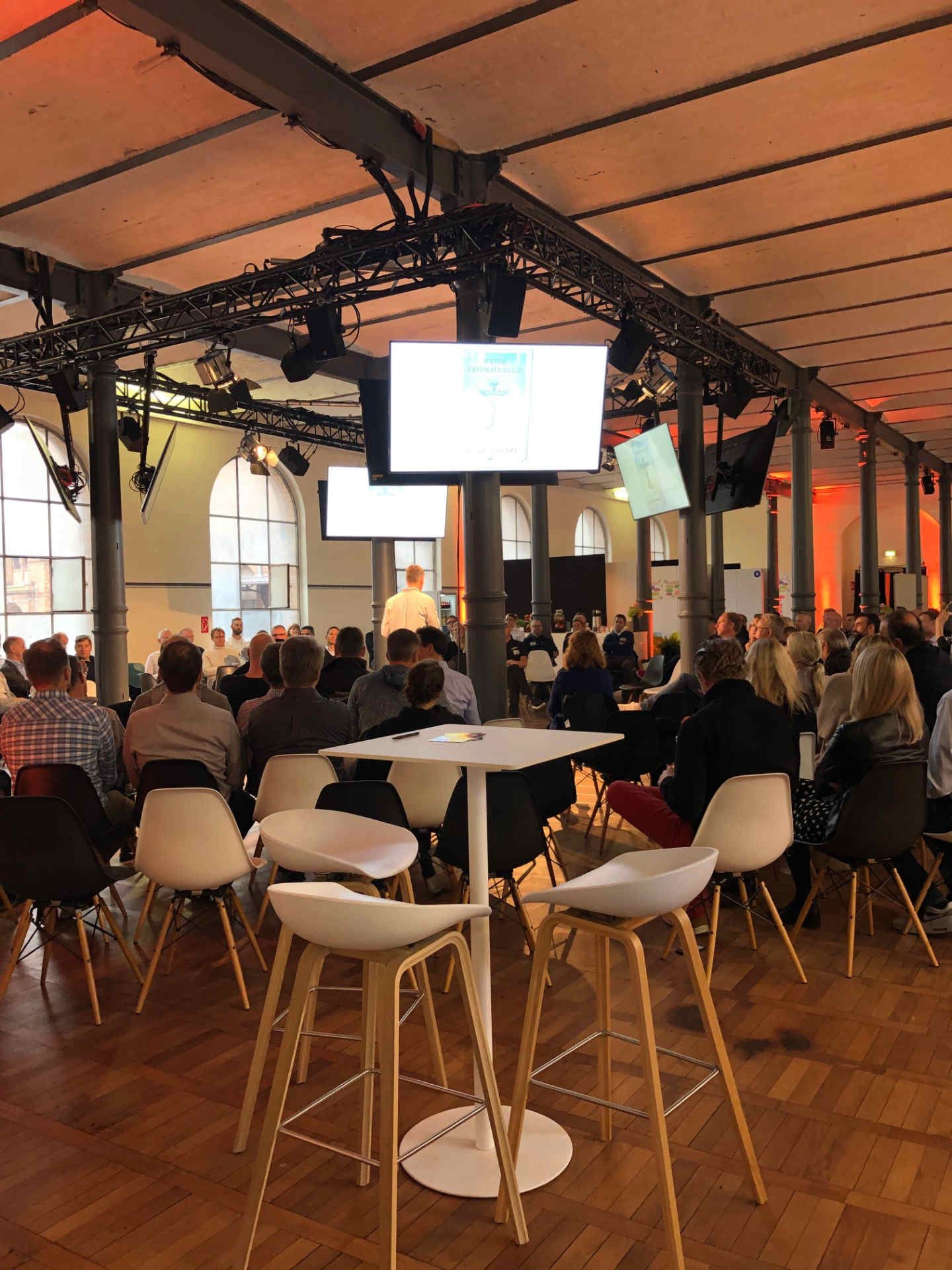 Eventsaal mit vielen Menschen zur Bühne schauend und Monitoren an Säulen