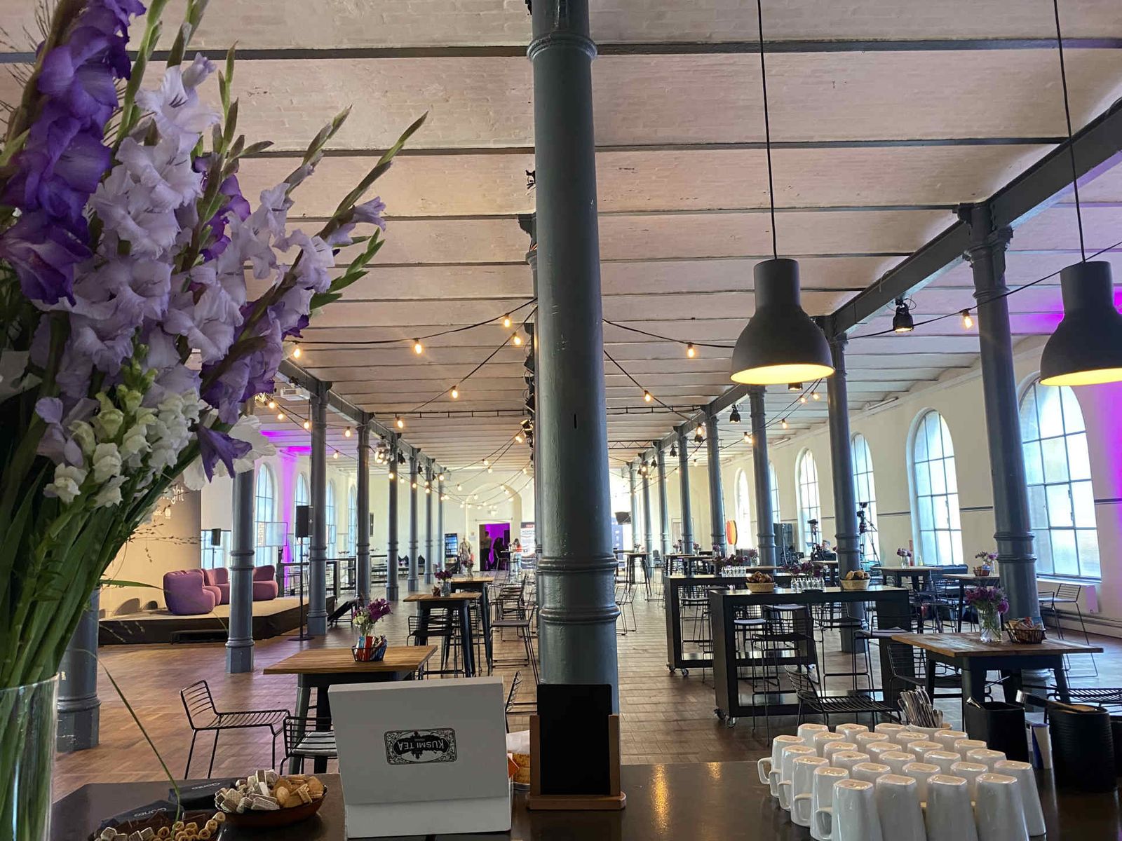 Großer Saal mit Blumendekoration und vielen Stehtischbrücken und einer Bühne im Hintergrund