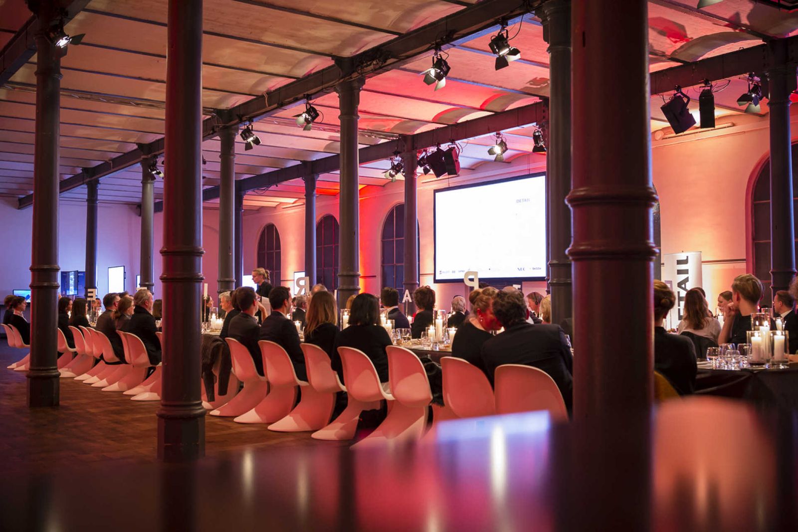 Langer Saal mit Säulen und langer Tafel an der Menschen auf Designer-Stühlen sitzen.