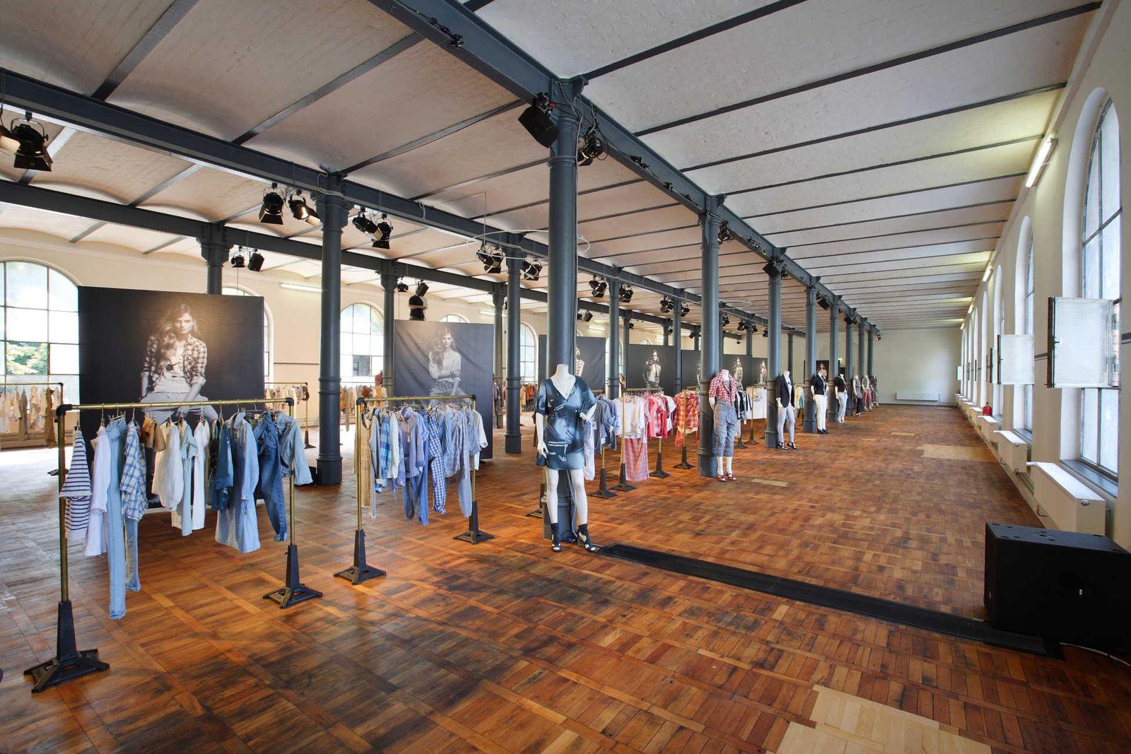 Long industrial hall with many clothing racks, mannequins and large posters.