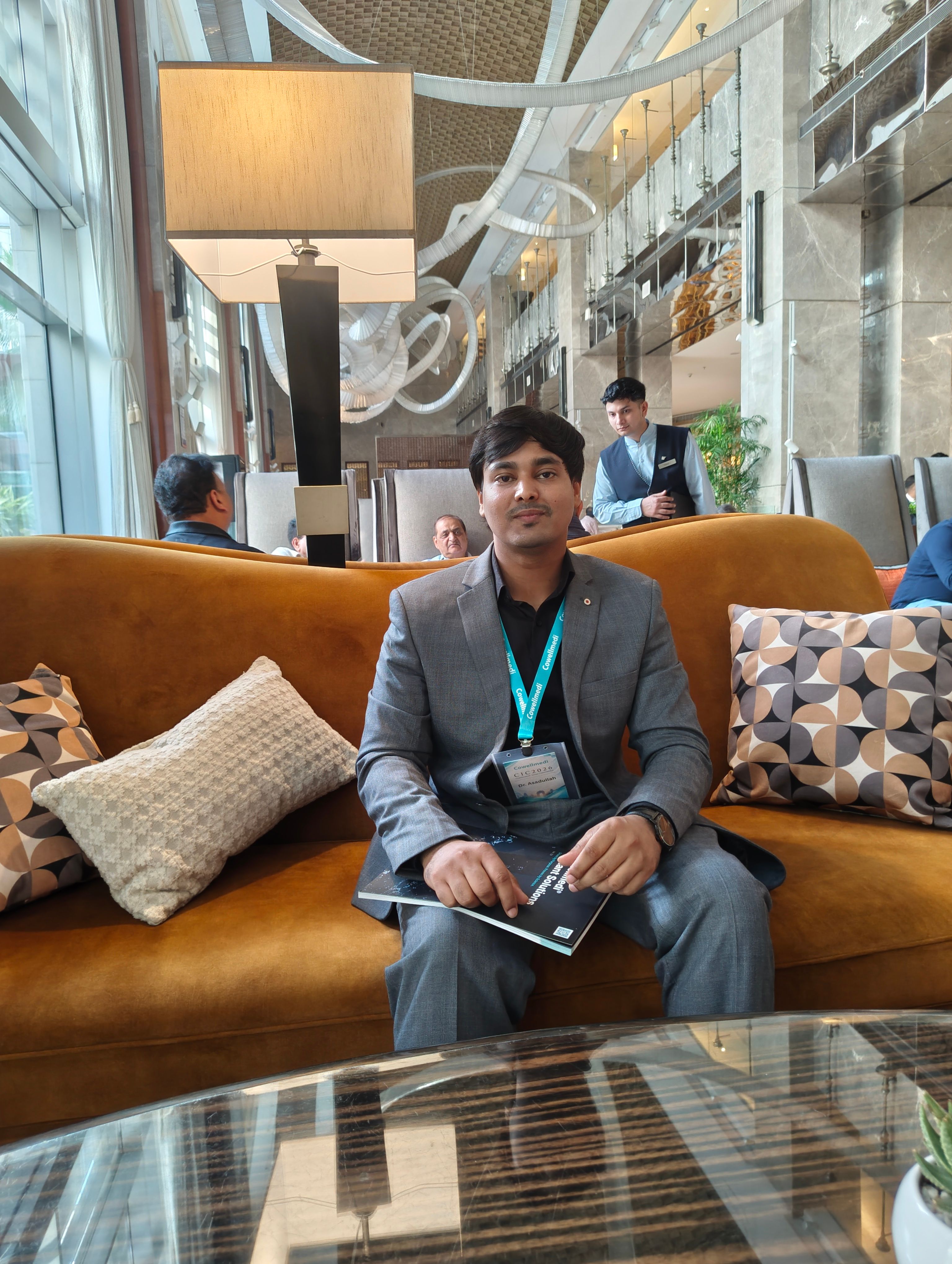 Dr Asad Frost seated on a mustard-gold sofa in an elegant hotel lounge, wearing a grey suit and black shirt, with books and a journal on the table in front of him.