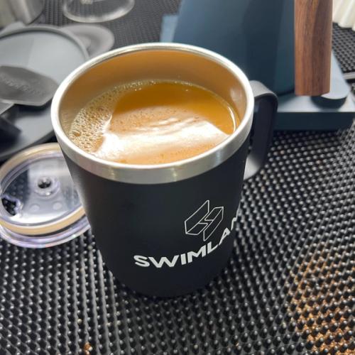 black tall metal mug with the text "swimlane" containing coffee with a splash of cream sitting ontop of a black bar mat amongst other coffee equipment