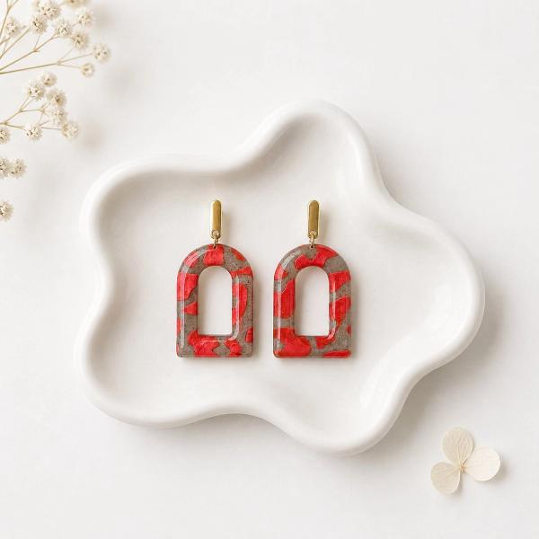 A pair of arch-shaped clay dangle earrings with a red and taupe terrazzo pattern, hanging from gold bar posts, displayed on a white ceramic tray.