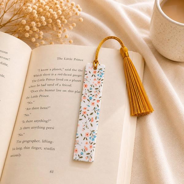A slim white bookmark printed with a ditsy pink and blue floral pattern and olive green leaves, with a gold braided tassel at the top, resting on an open book.
