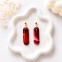 A pair of elongated pill-shaped dangle earrings with red and burgundy marble swirl pattern, hanging from small gold ball studs, displayed on a white wavy tray.